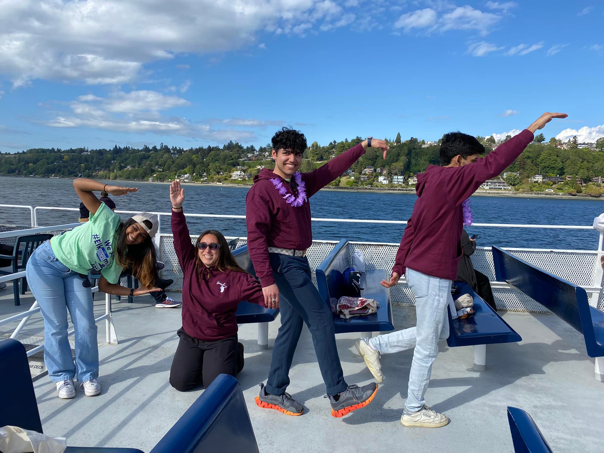 four Elks scholars stand on a boat with a Pacific Northwest scenery in the background, the scholars are spelling out E L K S