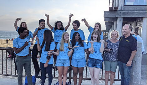 Elks scholars are standing and sitting in front of the beach. They're wearing a bright blue shirt that says 'Elks Scholar Service Trip.' They are pointing at their Elks Family