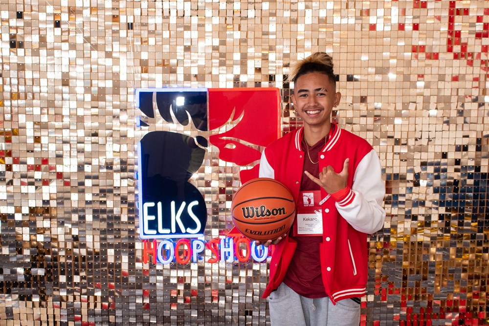 Kekane (a national finalist) stands in front of a shimmer backdrop with a neon hoop shoot logo while holding a basketball and showing a hang ten with his hand