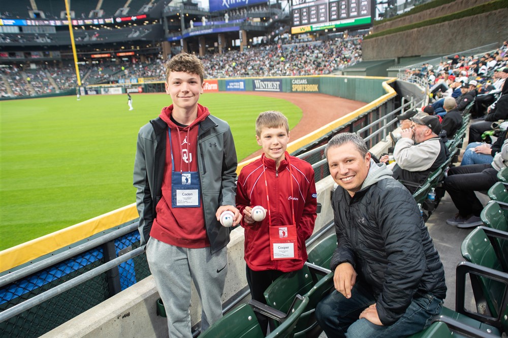 the Thompson family stands at Guaranteed Rate Field with the White Sox field behind them. Kaden and Cooper holding baseballs