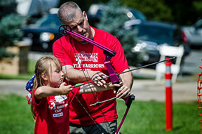 picture of man helping young girl shoot a bow and arrow
